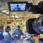 Close-up van een professionele videocamera gericht op chirurgen, met live weergave van de operatie op een monitor.